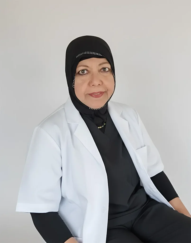 A woman in a white lab coat and black attire, wearing a black hijab, seated against a light background.