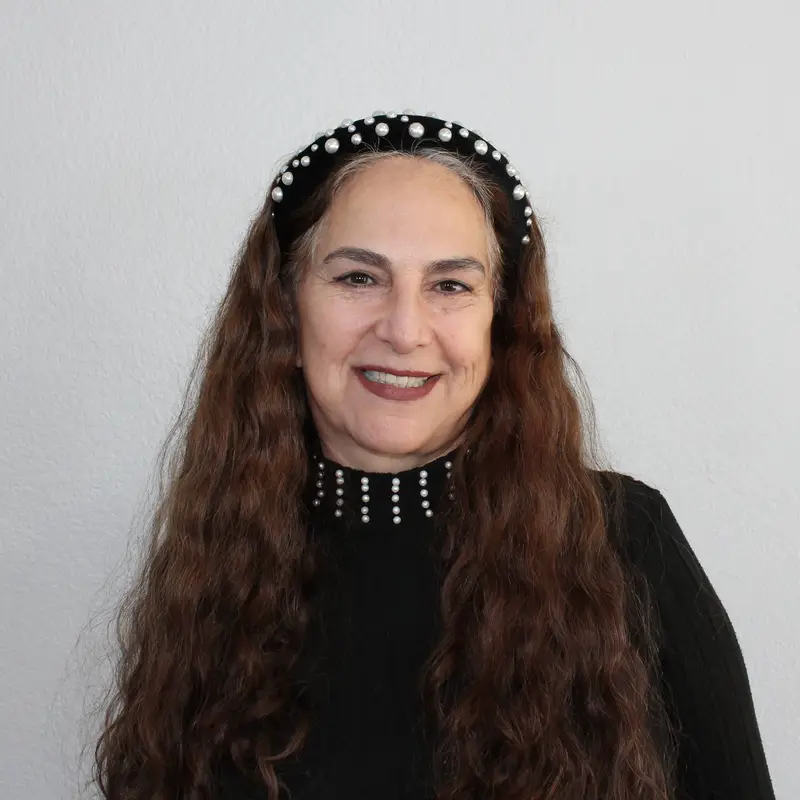 A woman with long wavy brown hair smiles at the camera. She is wearing a black top with pearl accents on the collar and a matching black headband with pearls. The background is plain and light-colored.