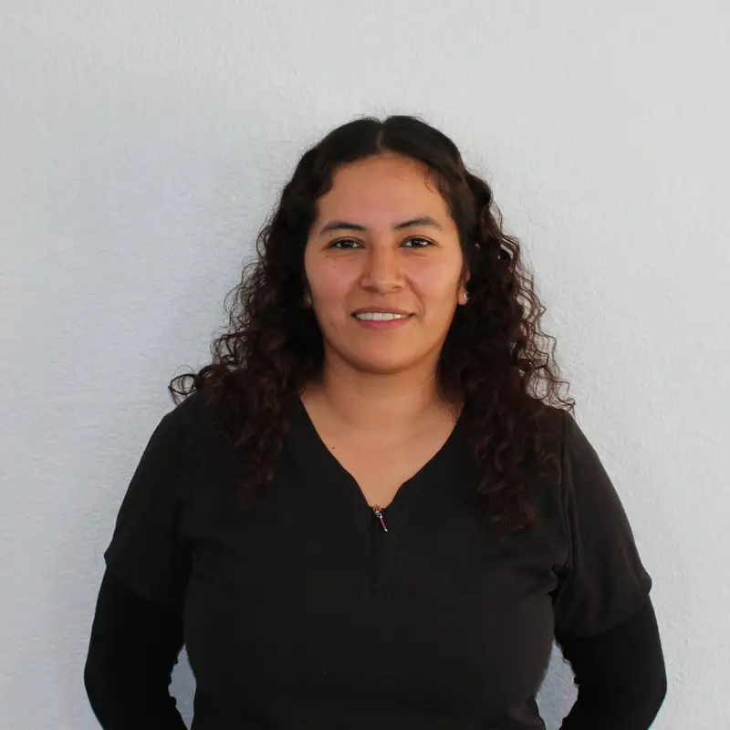 A woman with curly dark hair, wearing a black top, stands in front of a plain light-colored wall, looking at the camera and smiling gently.