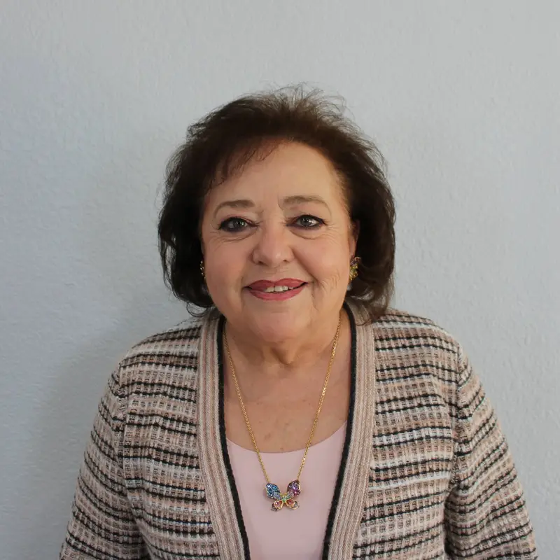 A smiling older woman with short brown hair, wearing a striped cardigan over a light pink top and a gold necklace with colorful stones, stands in front of a plain light gray wall.