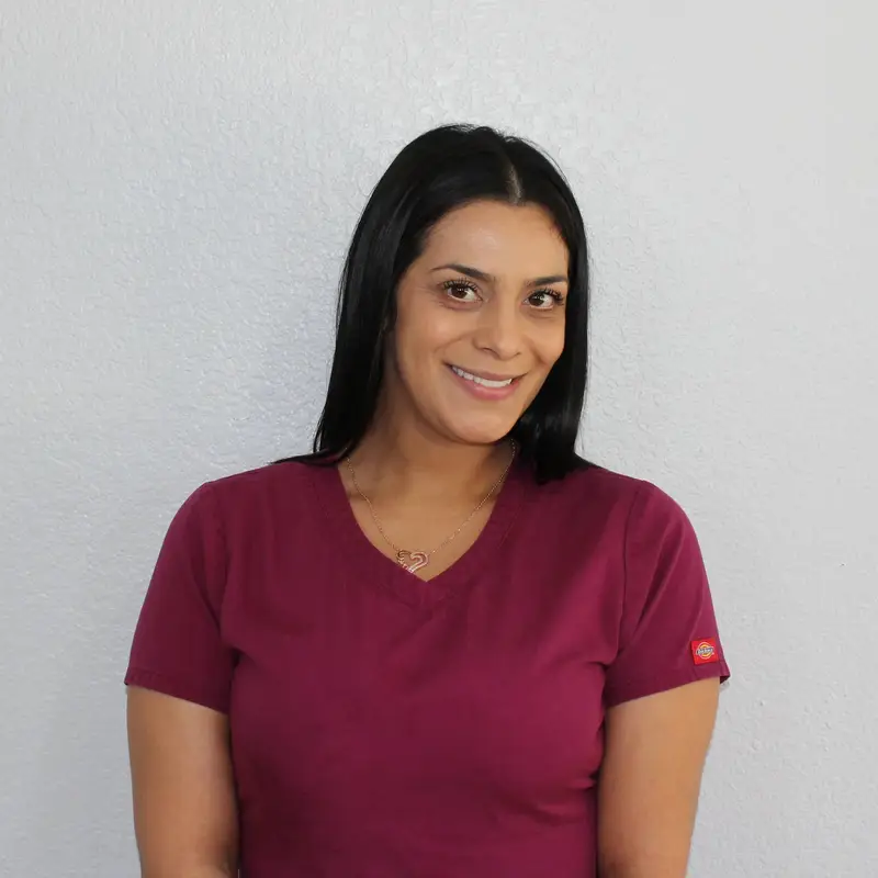 A woman with straight dark hair wearing a maroon scrub top stands in front of a plain light gray wall, smiling with her hands relaxed in front of her.