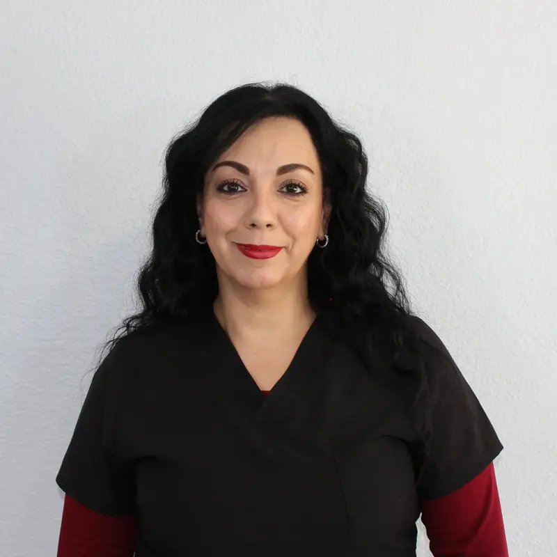 A woman with long, wavy black hair wearing a black scrub top over a red shirt stands against a plain light-colored wall, smiling slightly at the camera.