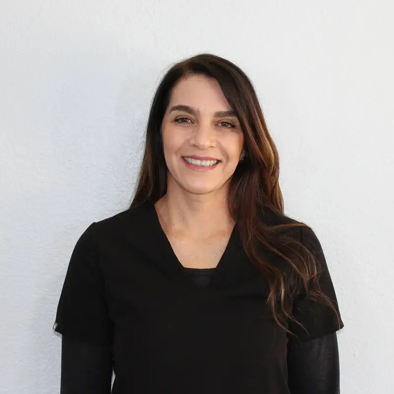A woman with long brown hair wearing a black top is standing and smiling in front of a plain white background.